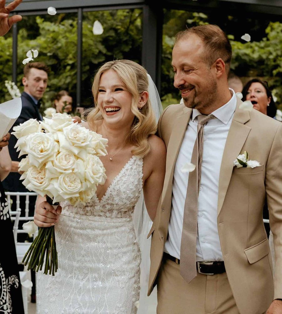 Glückliches Brautpaar bei Hochzeit, umgeben von Freunden, mit Blumen und Konfetti im Hintergrund.